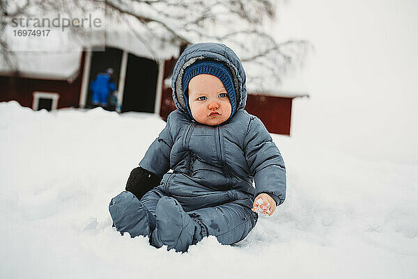 Niedliches Baby sitzt auf Schnee und berührt zum ersten Mal Schnee auf dem Bauernhof