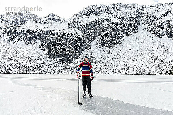 Ganzkörperlicher junger Mann mit Schläger und Puck  der lächelt und in die Kamera schaut  während er auf dem Eis eines gefrorenen Sees in der Nähe eines verschneiten Berges während eines Eishockeyspiels in der Landschaft von British Columbia  Kanada  steht