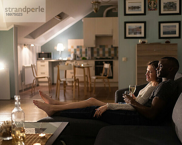 Verschiedenes Paar entspannt sich auf dem Sofa mit Wein am Abend