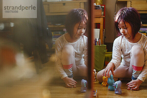 Ein kleines Kind sitzt gespiegelt im Glas und spielt mit Spielzeug auf dem Holzboden