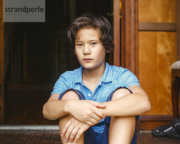 Ein Junge mit goldener Haut und ernster Miene sitzt in der Tür seines Hauses