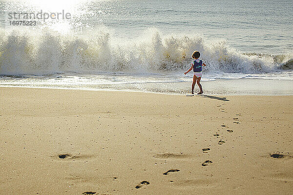 Ein kleines Mädchen in Schwimmweste steht vor einer entgegenkommenden Welle am Strand