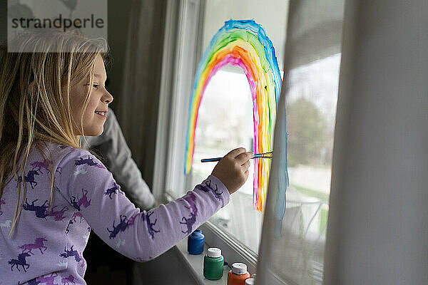 Seitenansicht eines lächelnden Mädchens  das einen Regenbogen auf ein Fenster malt