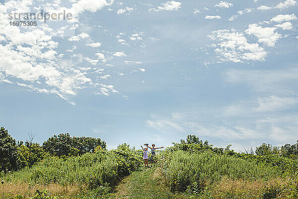 Zwei glückliche Kinder stehen mit ausgebreiteten Armen auf einem Hügel unter einem blauen Himmel