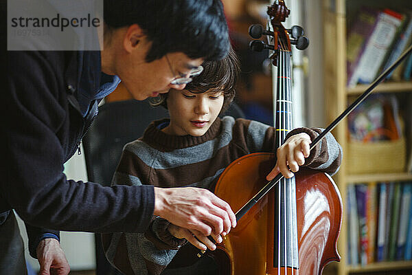Ein Vater beugt sich über sein Kind und hilft ihm  mit einem Bogen Cello spielen zu lernen