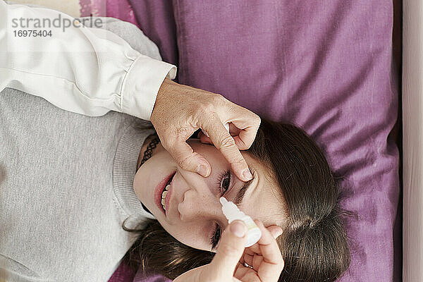 Draufsicht auf ein Detail einer lächelnden Ärztin  die einem kleinen Mädchen im Bett Augentropfen gibt. Hausarzt Konzept