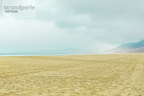 Unendlicher Strand mit Bergen im Hintergrund an einem dunstigen und bewölkten Tag. Strand von Cofete  Kanarische Inseln.