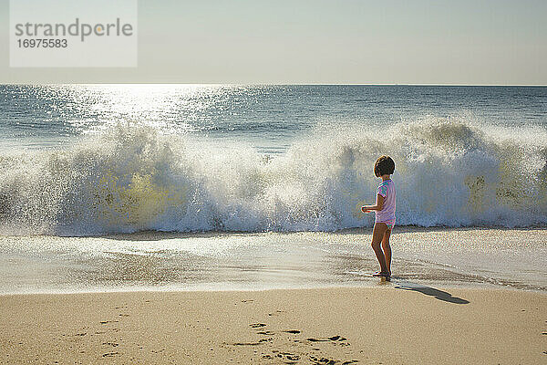 Ein kleines Mädchen steht am Rande des Ozeans und beobachtet  wie sich die Wellen am Strand brechen