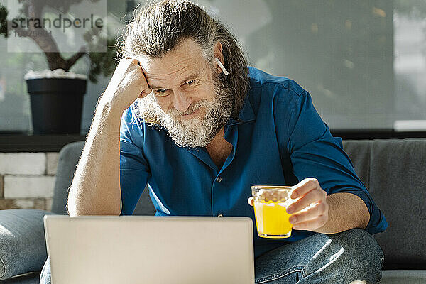 Älterer Mann mit Bart trinkt Orangensaft und schaut auf seinen Laptop  der auf einem Sofa sitzt. Business-Konzept