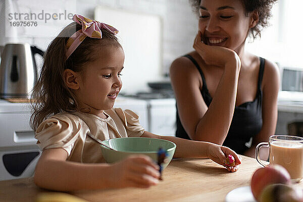 Fröhliches ethnisches Mädchen spielt beim Frühstück mit seiner Mutter