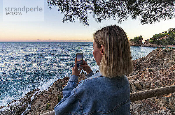 Junges blondes Mädchen beim Fotografieren des Sonnenuntergangs an der Küste