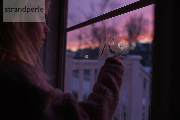 Kleines Mädchen schreibt auf Fensterbeschlag mit Sonnenuntergang im Hintergrund