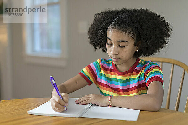 Zehnjähriges gemischtrassiges Mädchen schreibt am Tisch in ein Notizbuch