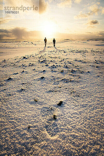 Zwei Menschen spazieren bei Sonnenaufgang am schwarzen Sandstrand in Island im Winter