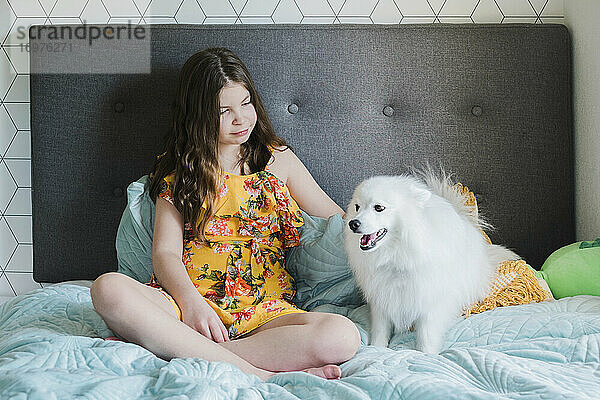 Junges Mädchen sitzt auf einem Bett und streichelt einen glücklichen kleinen flauschigen weißen Hund