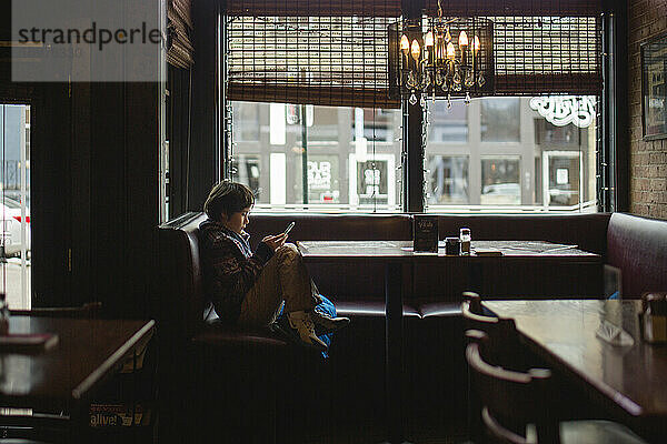 Ein kleiner Junge sitzt an einem Restauranttisch am großen Fenster und schaut auf sein Handy.