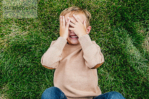 Glücklicher Junge lächelt und bedeckt sein Gesicht  während er im Gras liegt