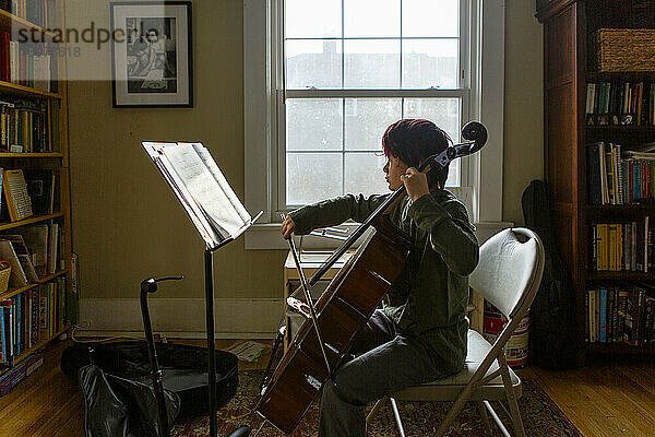Seitenansicht eines Jungen  der vor einem Fenster sitzt und in der Sonne Cello übt