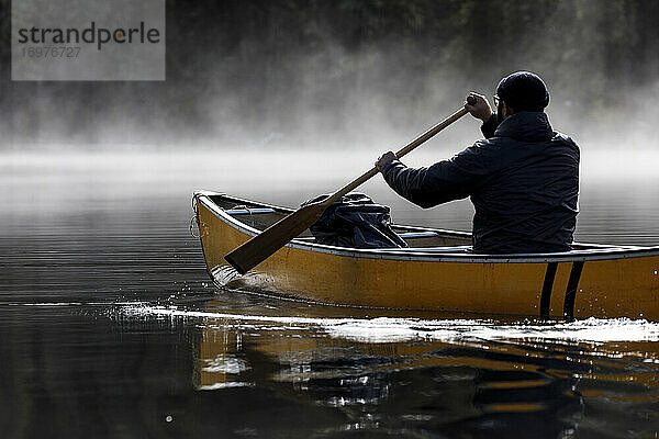 Konzentrierter Mann paddelt Kanu in den Nebel auf einem ruhigen See