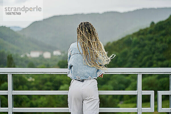 Rückseite einer jungen Frau mit blonden geflochtenen Haaren  die eine Jeansjacke und eine weiße Jeans trägt und sich an einem regnerischen Tag auf einem Damm ausruht