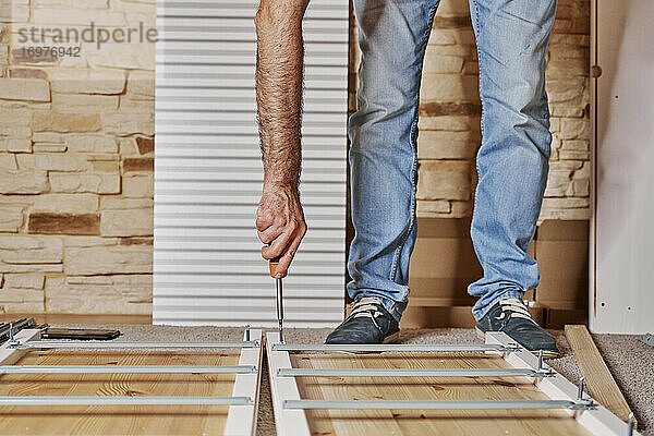 Vorderansicht eines unerkannten Arbeiters  der mit einem Schraubenzieher eine Schraube in eine Leiste eines Möbels schraubt  das er selbst aus Holz zusammenbaut. Horizontales Foto.