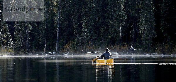 Mann paddelt Kanu auf einem See mit Nebel und Dunst an einem sonnigen Tag im Wald