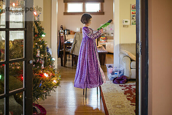 Ein kleines Kind in einem langen Prinzessinnenkleid steht auf einem Stuhl mit einer Spielzeugpistole
