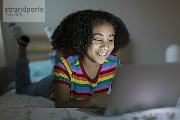 Lächelndes zehnjähriges gemischtrassiges Mädchen arbeitet an ihrem Apple-Laptop auf dem Bett