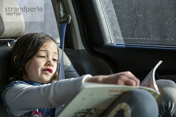 Ein süßes kleines Mädchen sitzt in einem Autositz in der Sonne und studiert ein Buch