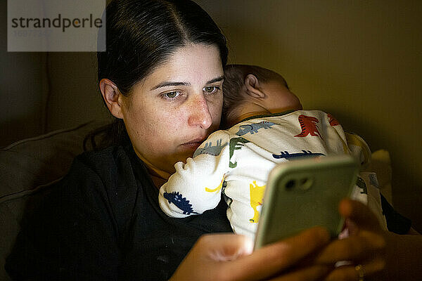 Mutter sitzt auf der Couch und surft im Internet auf einem Smartphone mit einem süßen schlafenden Baby in den Händen in einem dunklen Raum