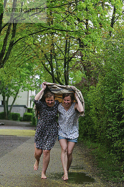 Zwei Mädchen laufen barfuß unter einem Regenmantel und lachen.