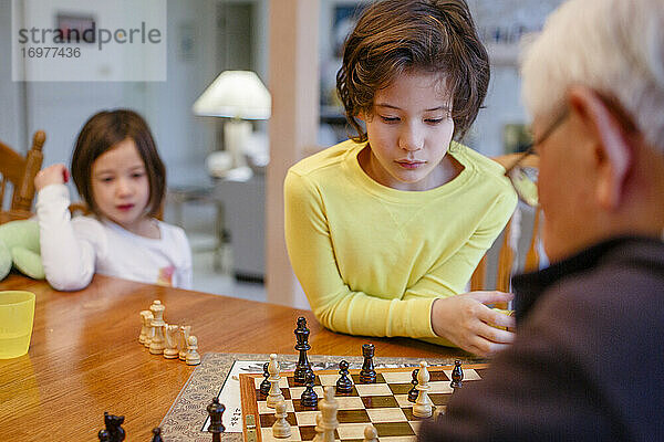 Ein Junge studiert ein Schachbrett  während seine Schwester und sein Großvater zuschauen