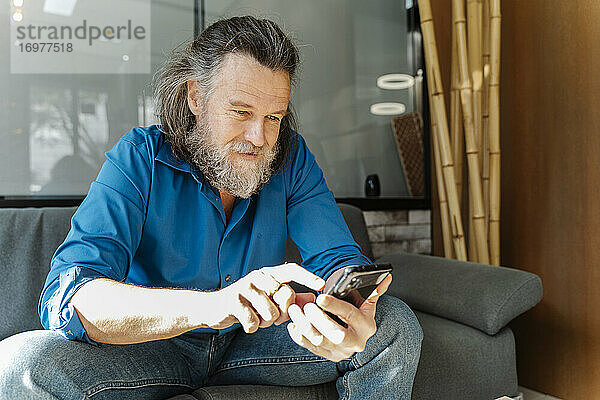 Älterer Mann mit Bart sitzt auf einem Sofa und schaut auf sein Smartphone im Wohnzimmer seines Hauses. Business-Konzept