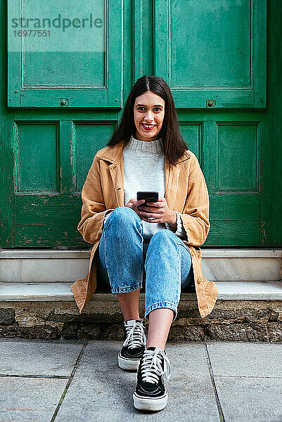 Porträt einer jungen stilvollen Frau  die im Freien sitzt und ein Smartphone benutzt