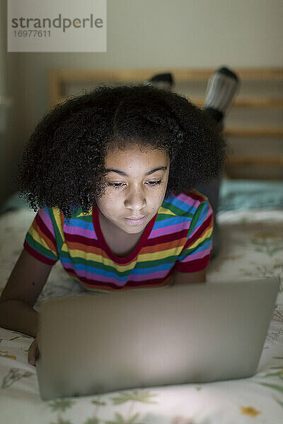 Zehnjähriges gemischtrassiges Mädchen arbeitet an ihrem Apple-Laptop  auf dem Bett liegend