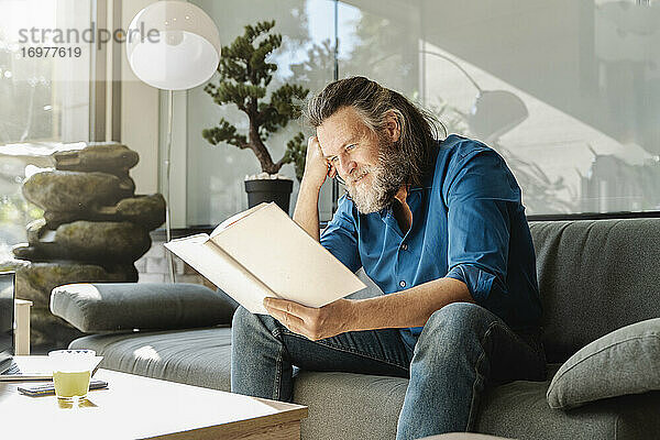 Älterer Mann mit Bart liest ein Buch auf dem Sofa im Wohnzimmer seines Hauses. Lernkonzept