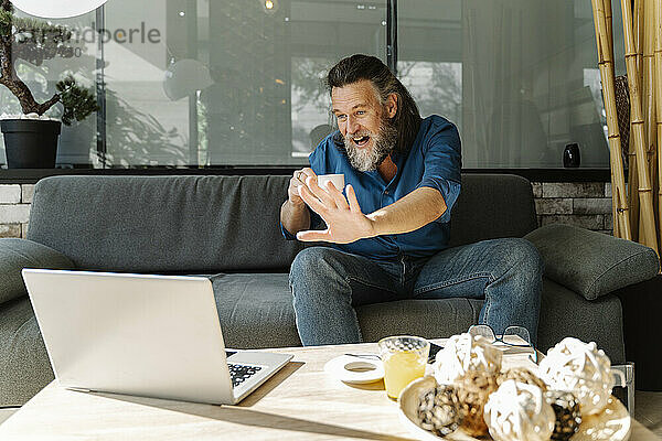 Älterer Mann mit Bart lächelt und arbeitet mit seinem Laptop auf einem Sofa zu Hause. Business-Konzept