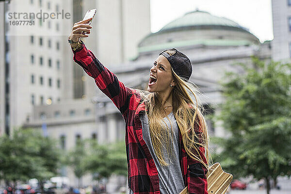 Stylische Städterin mit Skateboard macht ein Selfie in der Innenstadt