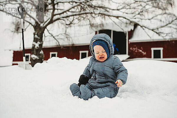 Weinendes Baby im Schnee sitzend in Norwegen mit roter Scheune dahinter