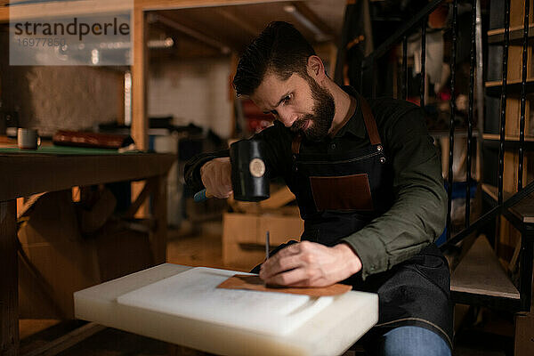 Bärtiger Handwerker bei der Herstellung von Lederhandwerk in einem gemütlichen Atelier