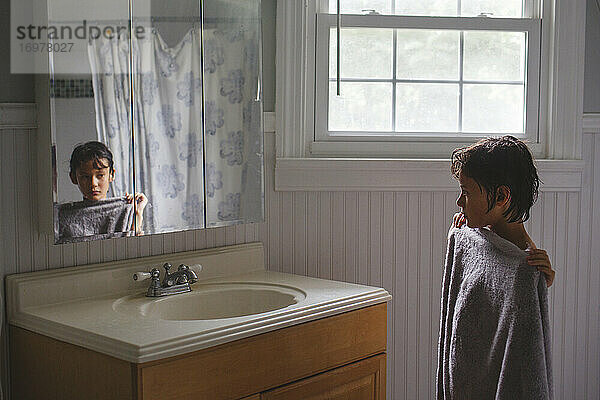 Ein Junge steht in ein Handtuch gewickelt vor der Dusche und schaut in einen Spiegel.