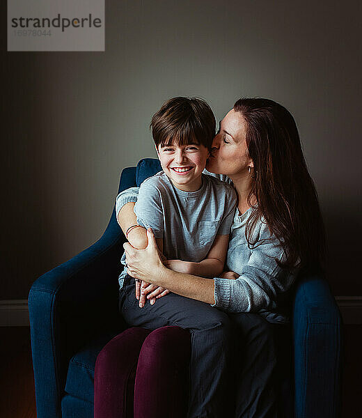 Eine Frau umarmt und küsst ihren Sohn  der in ihrem Schoß auf einem Stuhl sitzt.