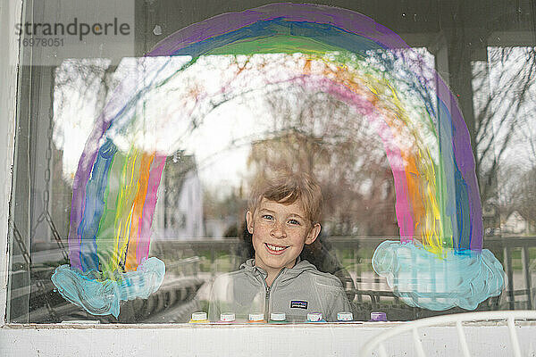 Blick von außen auf einen lächelnden Jungen unter einem auf das Fenster gemalten Regenbogen