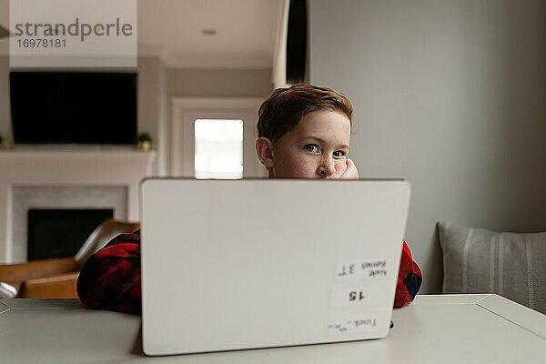 Junger rothaariger Junge blickt gelangweilt über einen Laptop-Computer drinnen am Tisch