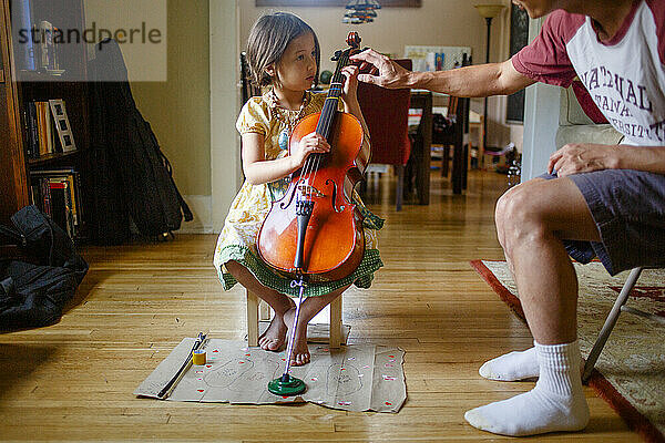Ein Vater beugt sich vor  um einem kleinen Mädchen beim Üben des Cellospiels zu helfen