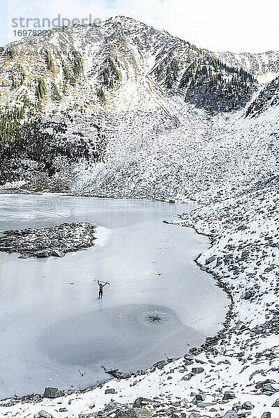 Drohnenansicht eines Sportlers  der auf einem zugefrorenen See in der Nähe eines verschneiten Bergrückens im Winter in der Natur von British Columbia  Kanada  Schlittschuh läuft und Hockey spielt