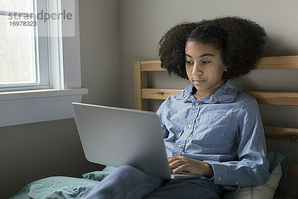 Zehnjähriges gemischtrassiges Mädchen arbeitet im Bett sitzend an ihrem Laptop