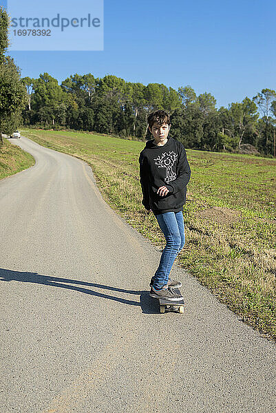 Junger männlicher Teenager mit Kapuze fährt auf einem Skateboard auf einer Bergstraße