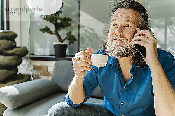 Älterer Mann trinkt einen Kaffee und telefoniert auf einem Sofa sitzend