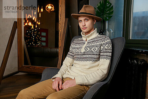 Porträt gut aussehend modischen Mann in einem weißen Pullover und braunen Hut sitzt auf einem Stuhl in einem Fotostudio Loft. Junger Geschäftsmann denkt über die Zukunft nach